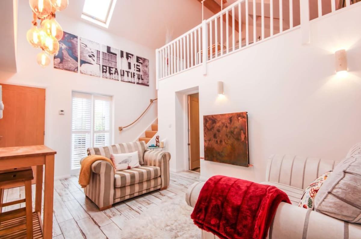 A bright open-plan living area features a comfortable striped sofa and a stylish armchair with colorful throws. Natural light pours in through the skylight and windows, highlighting the warm wood tones and artistic wall decorations, enhancing the cozy ambiance of the space.