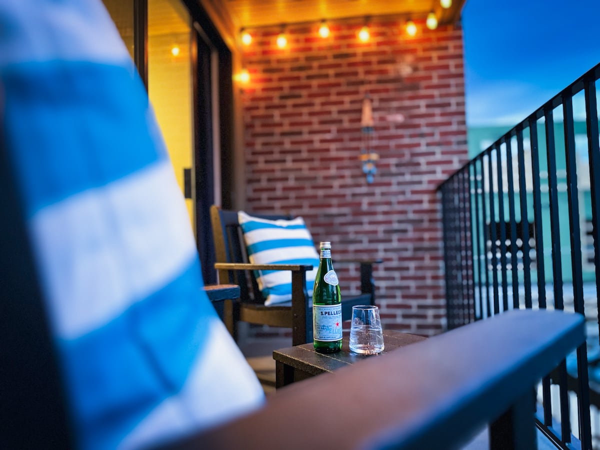 An inviting balcony scene features two wooden chairs adorned with striped cushions. A small table holds a glass of sparkling water and a bottle. Soft warm lights hang above, enhancing the cozy outdoor ambiance with a view of the surrounding area.