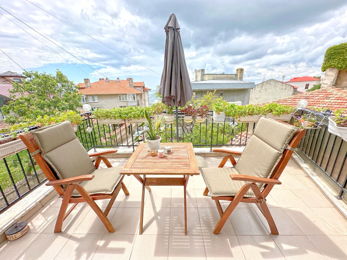 An outdoor terrace is adorned with two wooden chairs and a small table, surrounded by greenery. A sun umbrella provides shade, and a view of the neighboring rooftops is visible in the background under a partially cloudy sky.