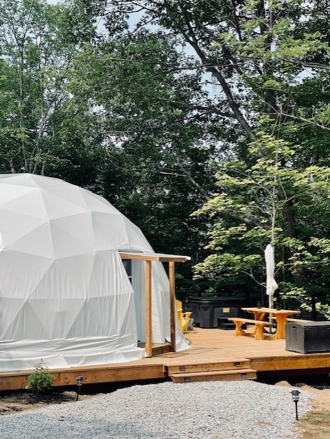 A white dome structure is nestled among greenery, showcasing a wooden deck with outdoor seating. The area features a picnic table and casual lounge furniture, surrounded by trees that provide a natural setting, ideal for relaxation.