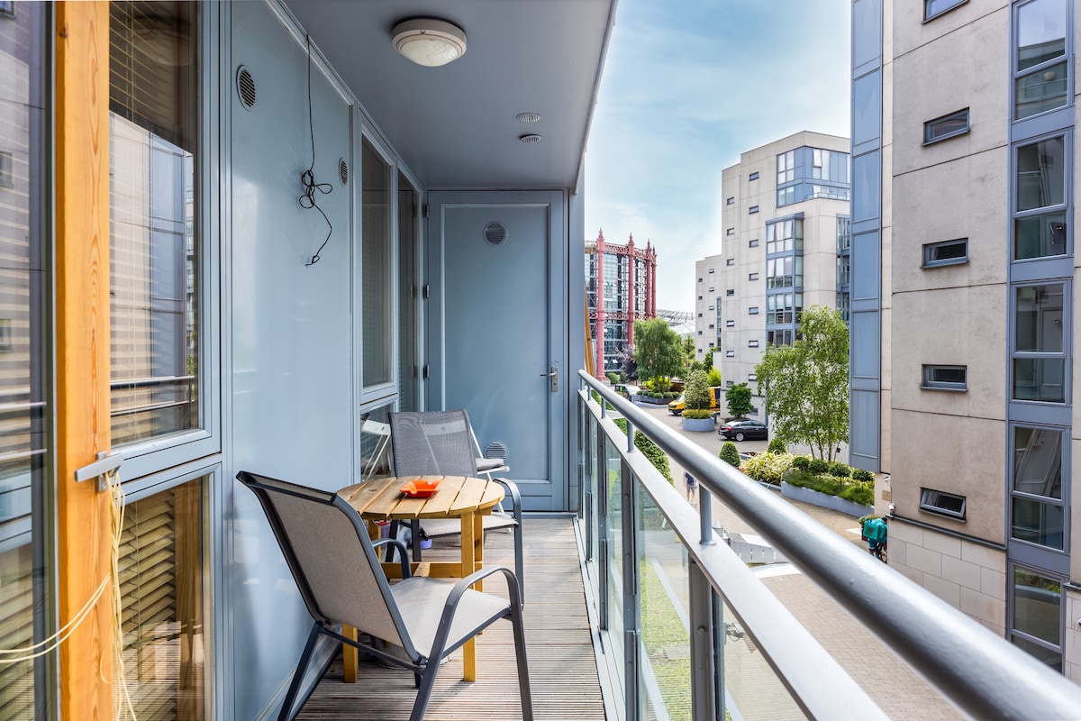 A spacious balcony features a wooden table and two chairs, providing a comfortable outdoor seating area. The view showcases the surrounding buildings and greenery, while natural light brightens the space. A glass railing ensures safety while enhancing visibility.