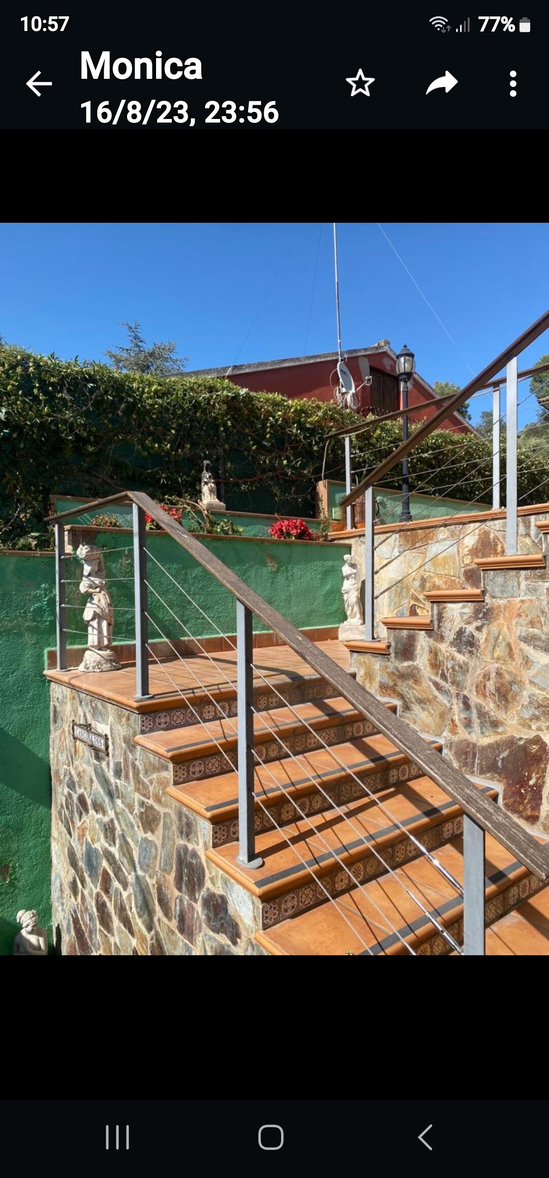 A stone staircase ascends with a railing featuring a modern design. Lush greenery is visible on both sides, enhancing the natural surroundings. Decorative statues are placed along the path, adding visual interest. Clear skies are seen above, contributing to an open and welcoming atmosphere.