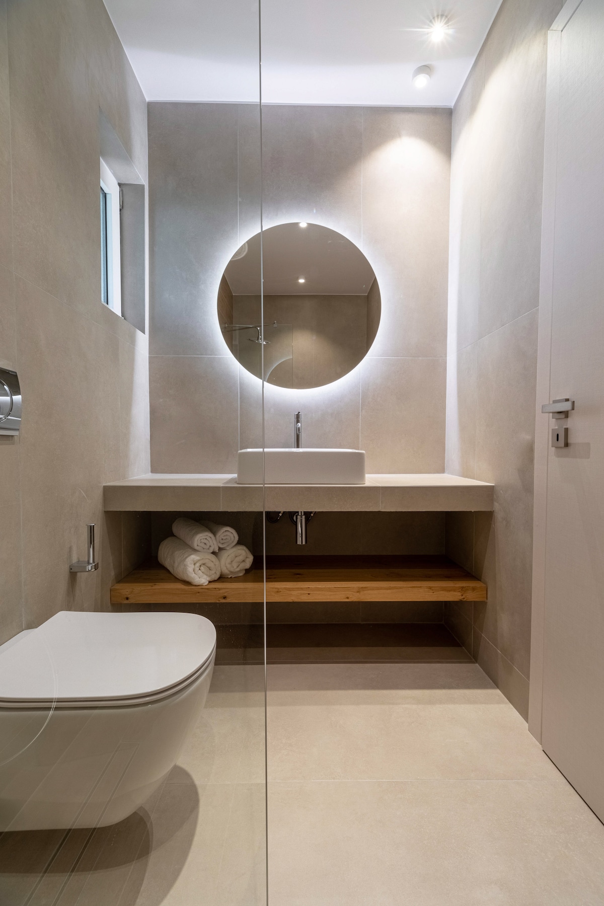 A modern bathroom features a minimalist design with light-colored tiles. A round mirror illuminated by soft lighting hangs above a sleek sink, while a wooden shelf underneath holds neatly rolled towels. A contemporary toilet is positioned nearby, and a small window allows for natural light.