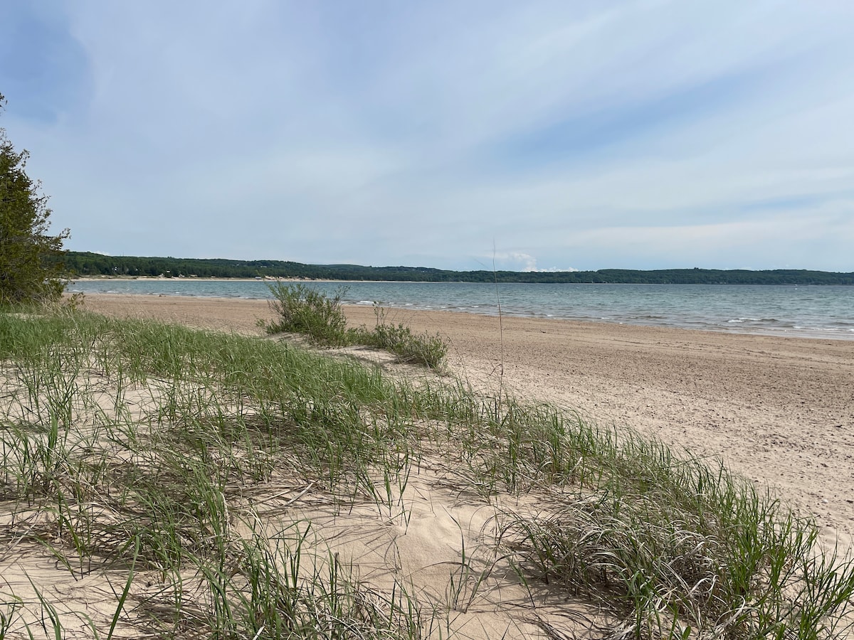 A sandy beach stretches along the shoreline of Little Traverse Bay, with gentle waves lapping at the water's edge. Grassy patches of dune vegetation are visible in the foreground, leading the eye toward the calm blue water and distant tree-lined horizon under a pale sky.
