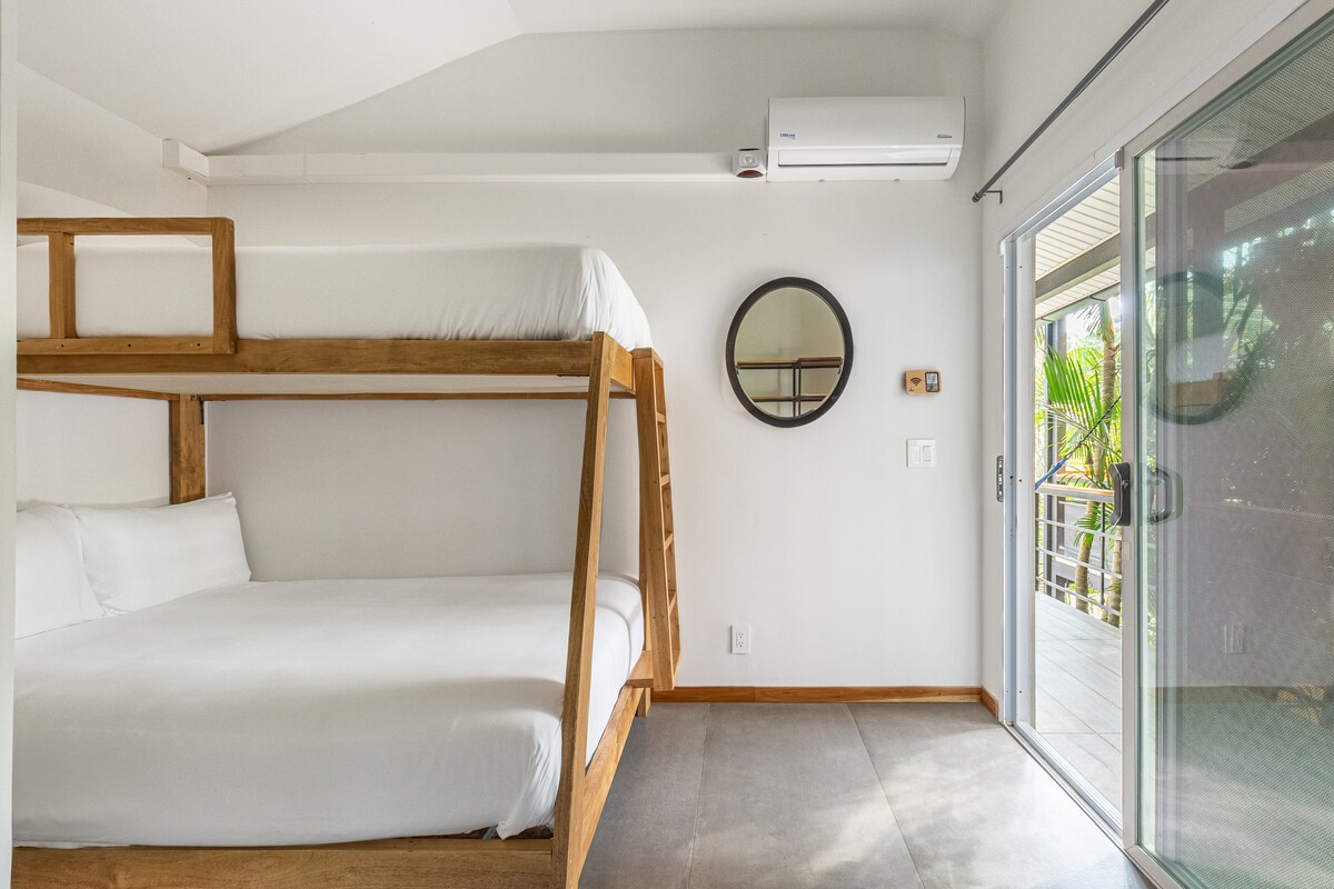 A bright room features a wooden bunk bed with a spacious lower sleeping area and a top bunk, both dressed in crisp white linens. A round mirror is mounted on the wall, while a glass door opens to an outdoor space, allowing natural light to fill the area.