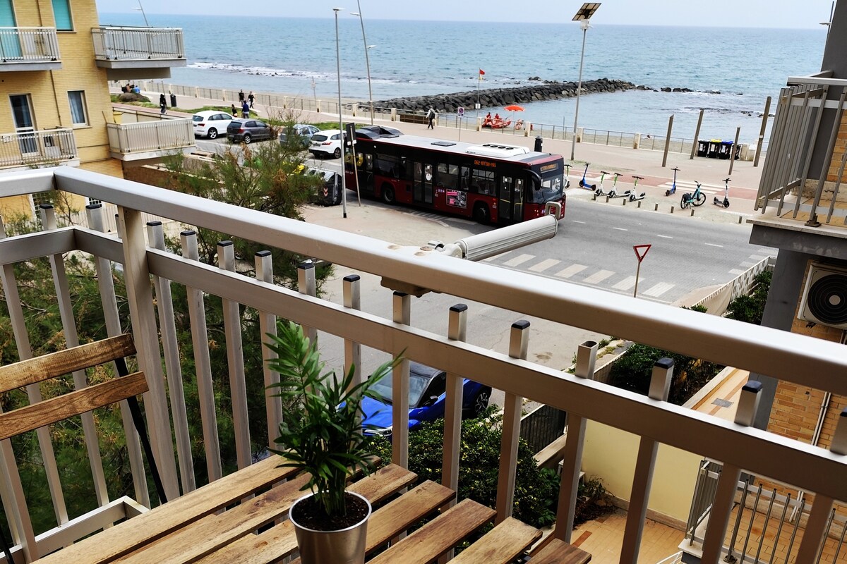 A view from a balcony features a small potted plant on a wooden table, overlooking a beach and the sea. A bus is seen on the street, with pedestrians walking along the coastline, and a pathway lined with bicycles in the foreground.