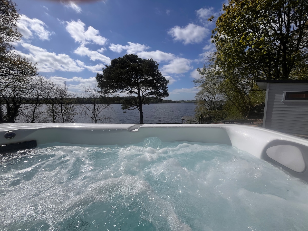 An outdoor hot tub is visible, featuring bubbling water, with a scenic view of the lake in the background. Lush trees frame the image, and a partly cloudy sky allows for light to reflect on the water's surface.