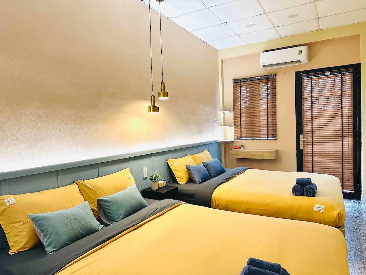 A cozy bedroom features two spacious beds dressed in cheerful yellow linens, complemented by soft blue accent pillows. Stylish pendant lights hang above the beds. Natural light filters through wooden blinds, illuminating a small shelf with a decorative plant.