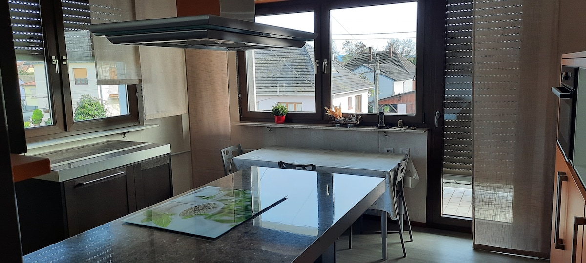A modern kitchen area features a central island with a smooth countertop and seating for two. Large windows provide natural light, showcasing views of the neighborhood. Kitchen appliances, including an oven and a cooktop, are visible, with light-colored cabinetry enhancing the spacious feel.