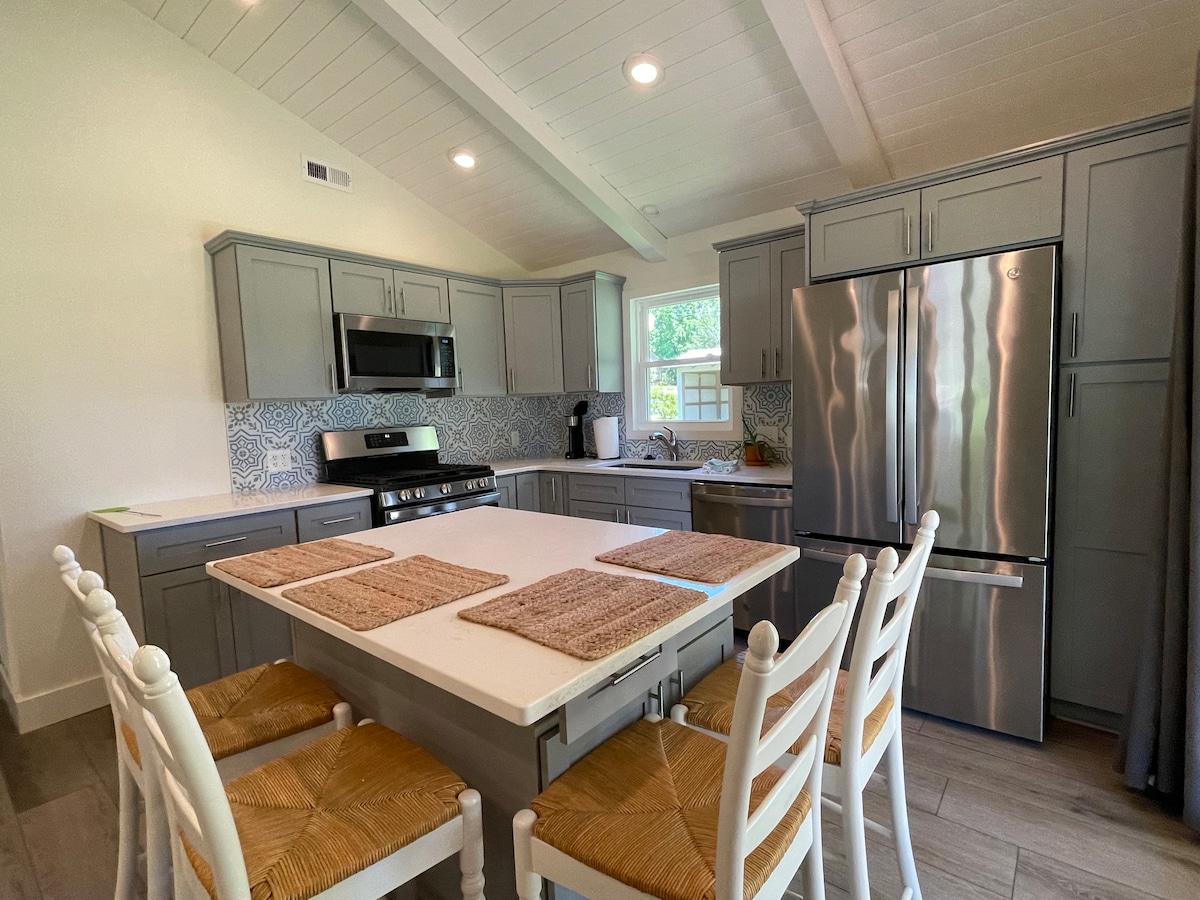 A modern kitchen is presented with gray cabinetry and stainless steel appliances, including a refrigerator and stove. An island with a white countertop offers seating for six on rustic chairs. Textured placemats are arranged on the table, completing the inviting space.