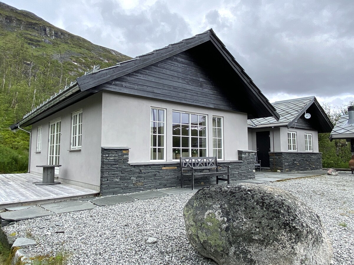A modern cabin is presented with a combination of stone and wood exteriors, featuring large windows that allow natural light to fill the interior. A gravel path leads to the entrance, with a wooden bench situated in front of the house.
