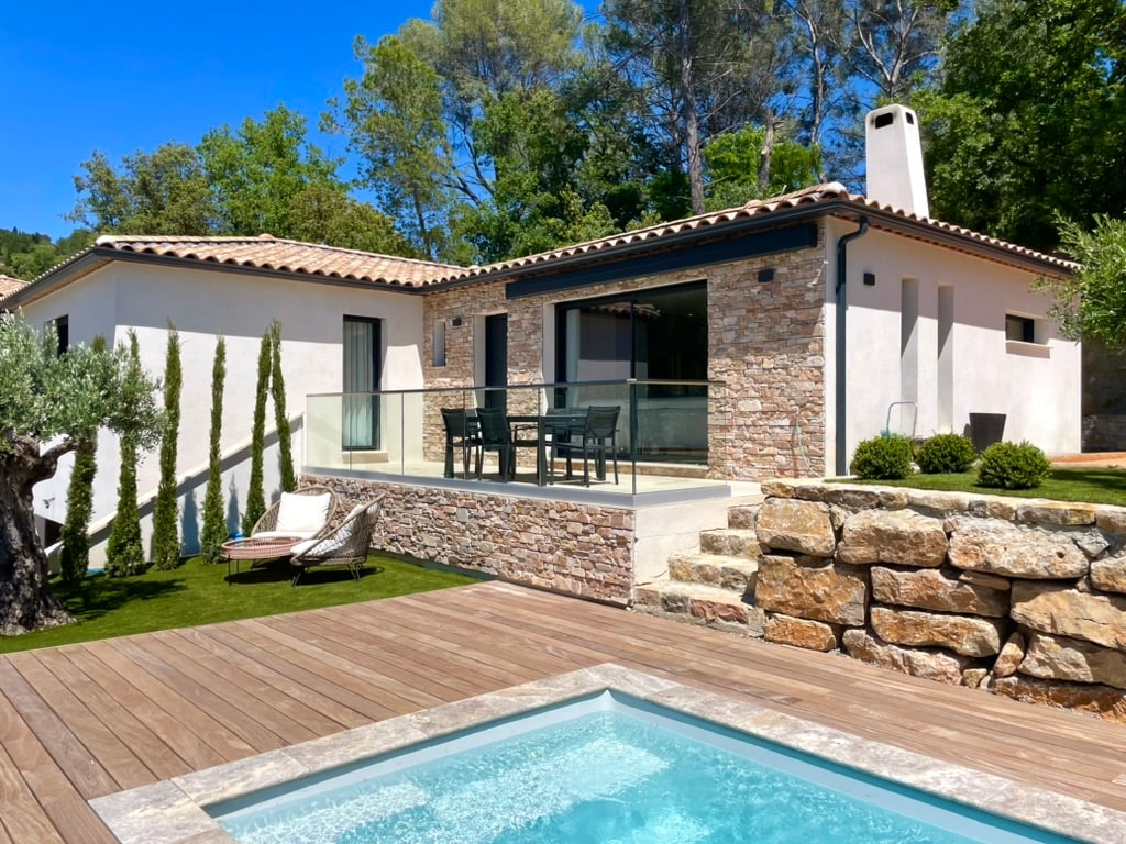 The villa's exterior is showcased with a stone facade and large glass doors leading to a terrace. A private swimming pool glimmers nearby. Lush greenery and neatly trimmed trees enhance the outdoor setting, while a comfortable lounge chair is positioned on the wooden deck.