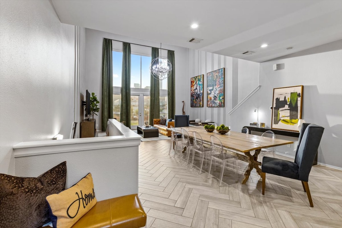 A spacious living and dining area features large windows framed by floor-length curtains, allowing natural light to fill the room. A modern wooden dining table is surrounded by transparent chairs. Artwork adorns the walls, while a comfortable seating arrangement is visible in the background.