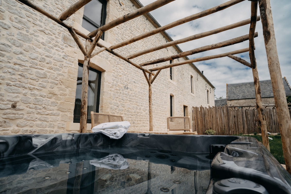 A hot tub is outlined by a rustic wooden structure, situated in a serene outdoor area. The natural stone building features large windows, allowing ample light. Two wicker chairs provide seating, creating a comfortable space for relaxation. A towel rests neatly on the edge of the hot tub.