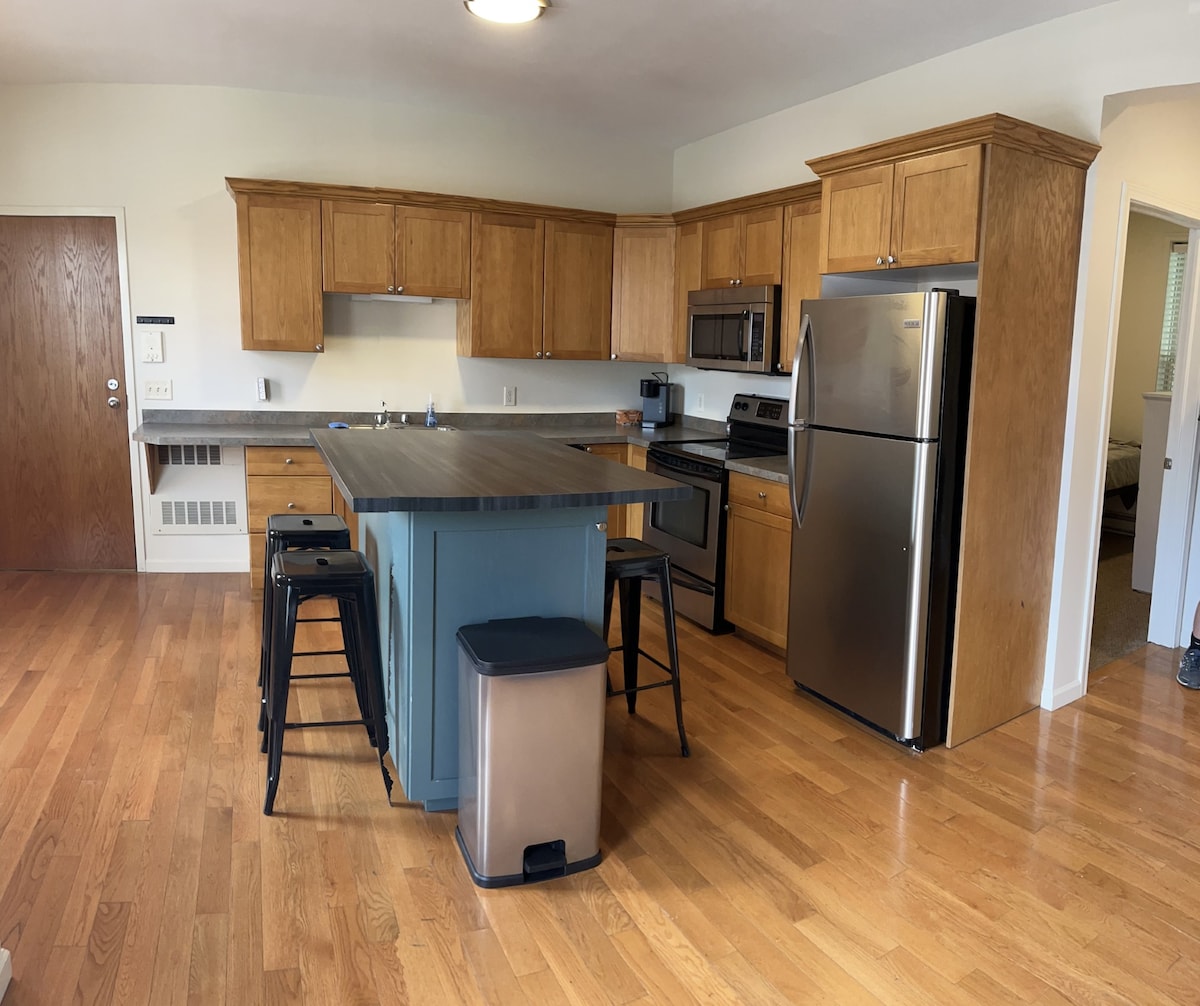 A modern kitchen showcases wooden cabinetry and stainless steel appliances. An island features a dark countertop with seating for four. Hardwood flooring extends throughout the space, creating a cohesive look. A clean and organized countertop is ready for meal preparation.