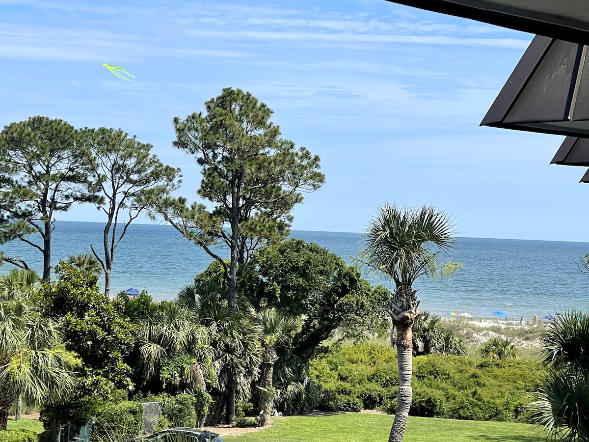 The image captures a peaceful ocean view framed by lush greenery and palm trees. The expansive blue sky meets the calm sea at the horizon. In the distance, beach umbrellas can be seen along the shore, enhancing the coastal ambiance.
