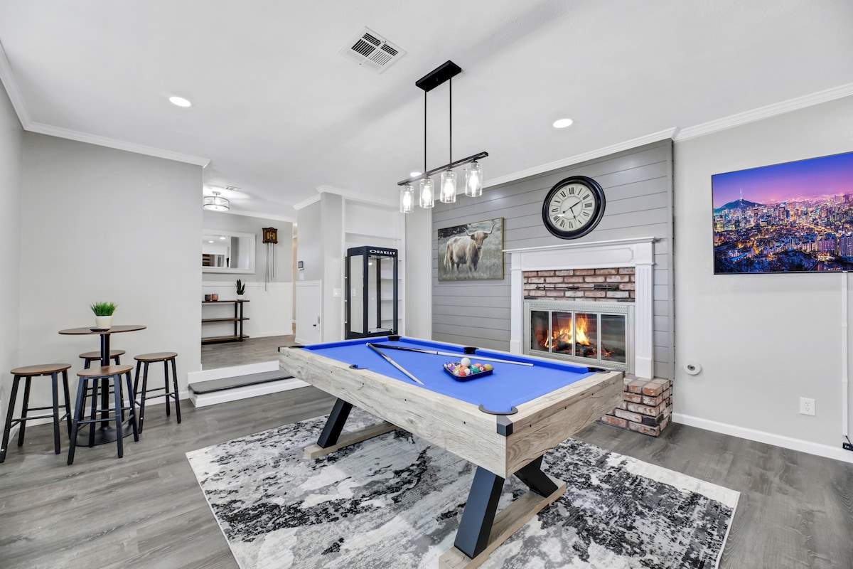 An open-concept game room features a blue pool table at its center, surrounded by a large area rug. A stone fireplace with a modern mantle and a mounted television displaying a cityscape adds warmth to the space, which is illuminated by stylish pendant lighting.