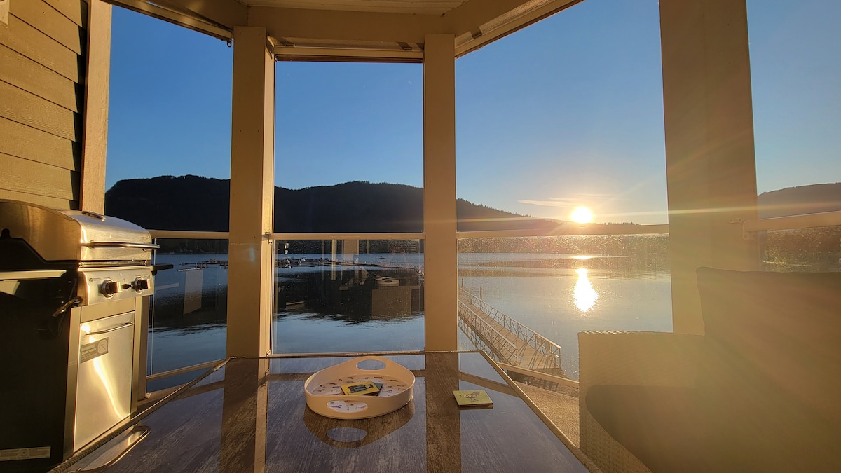 A spacious balcony is illuminated by the golden hues of a sunset. A gas grill is positioned to the left, while a table with a circular tray is placed in the center. The tranquil water reflects the scenic landscape, framed by hills in the distance.
