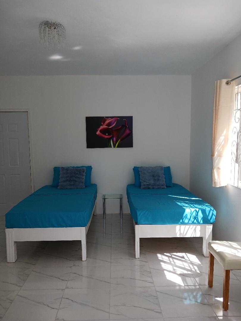 A bright room featuring two single beds dressed in vibrant blue covers. A small glass table is situated between the beds. Natural light filters through a nearby window, illuminating a floral artwork on the white wall. The floor is tiled in a glossy white finish.
