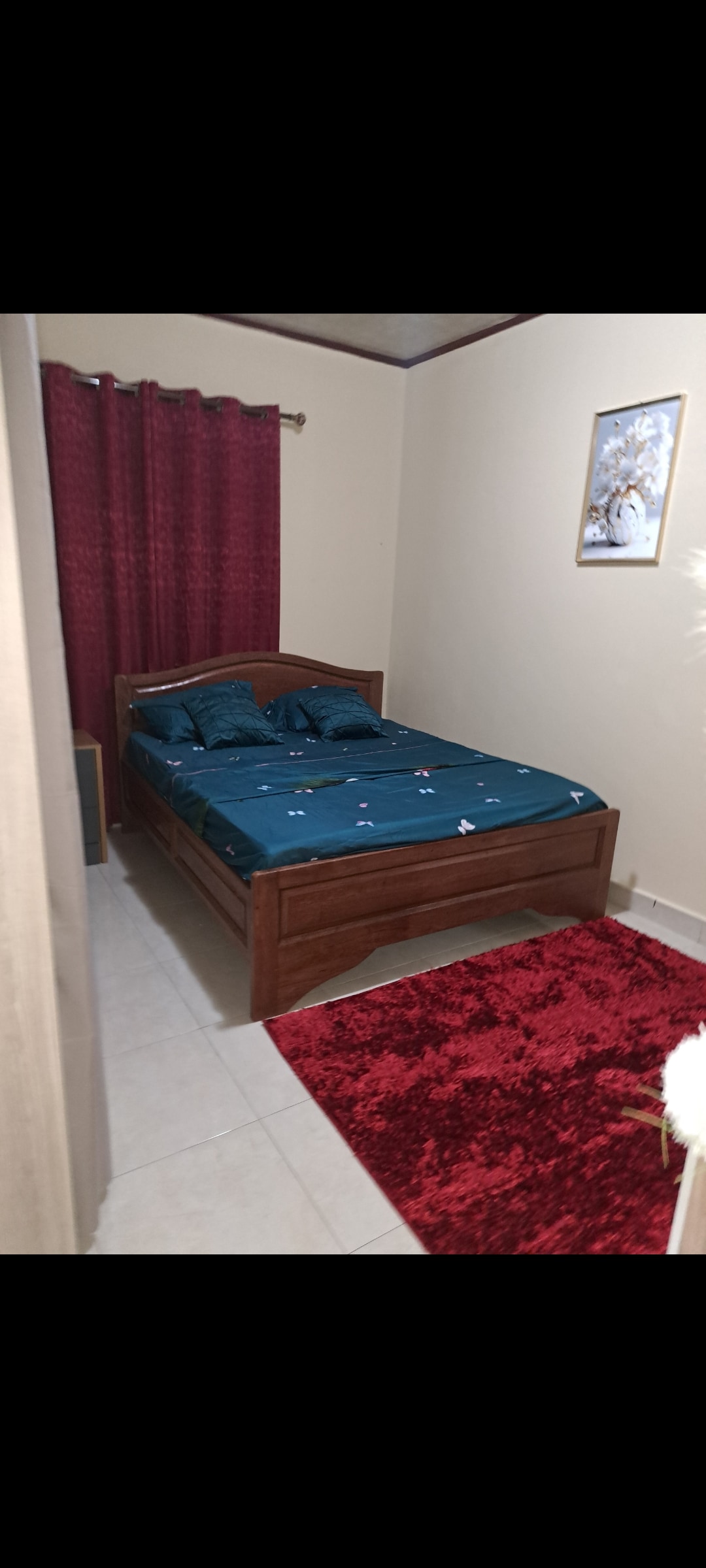 A simple bedroom is furnished with a wooden bed covered in a dark teal bedspread. Two decorative pillows are arranged on the bed. A deep red curtain frames the window, and a plush red area rug adds warmth to the floor. A framed piece of artwork is hung on the wall.