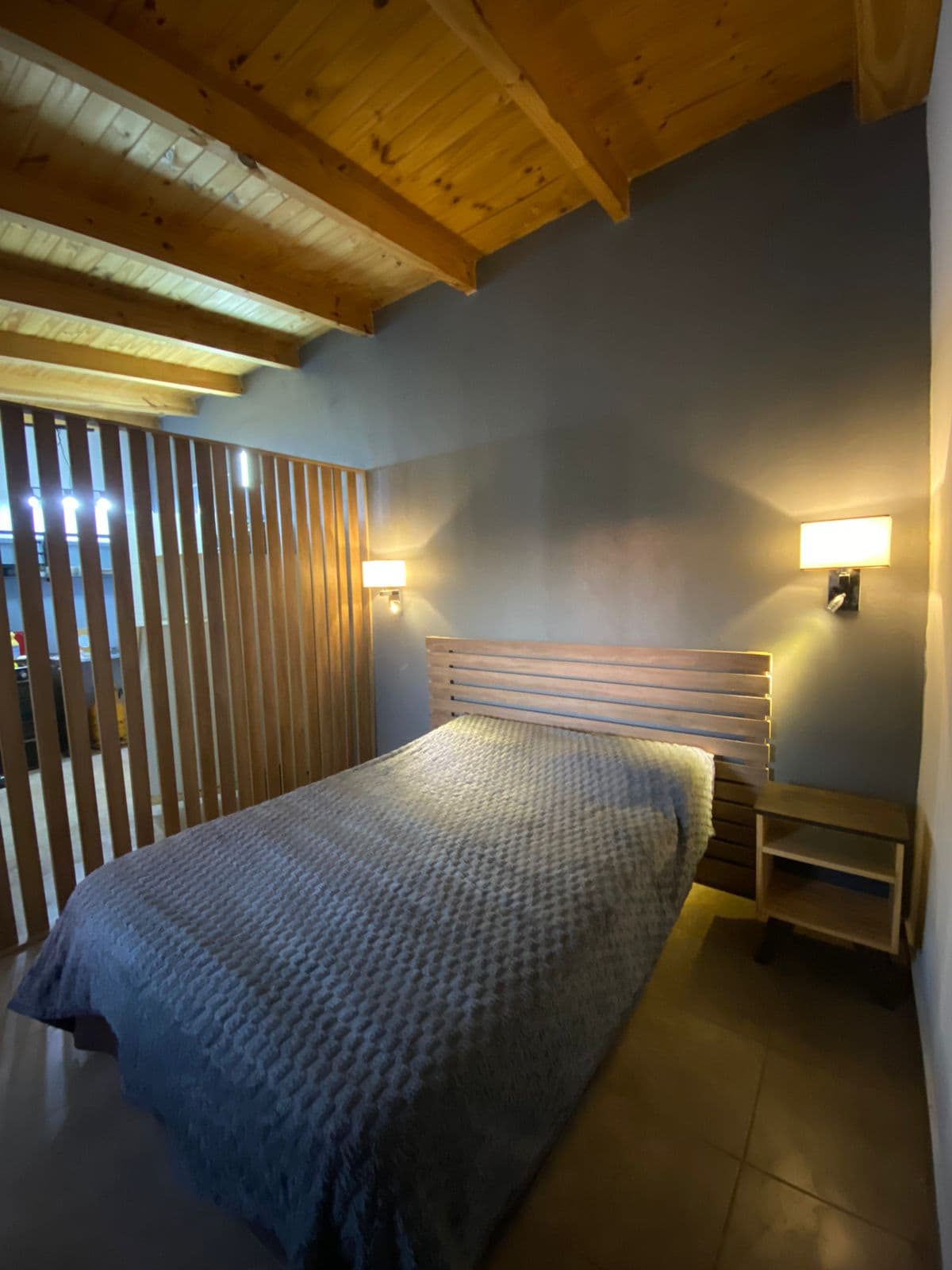 A cozy bedroom features a spacious bed with a textured gray coverlet. Soft lighting is provided by wall sconces on either side of the bed. A wooden slatted partition adds a sense of privacy, while exposed wooden beams enhance the natural feel of the space.