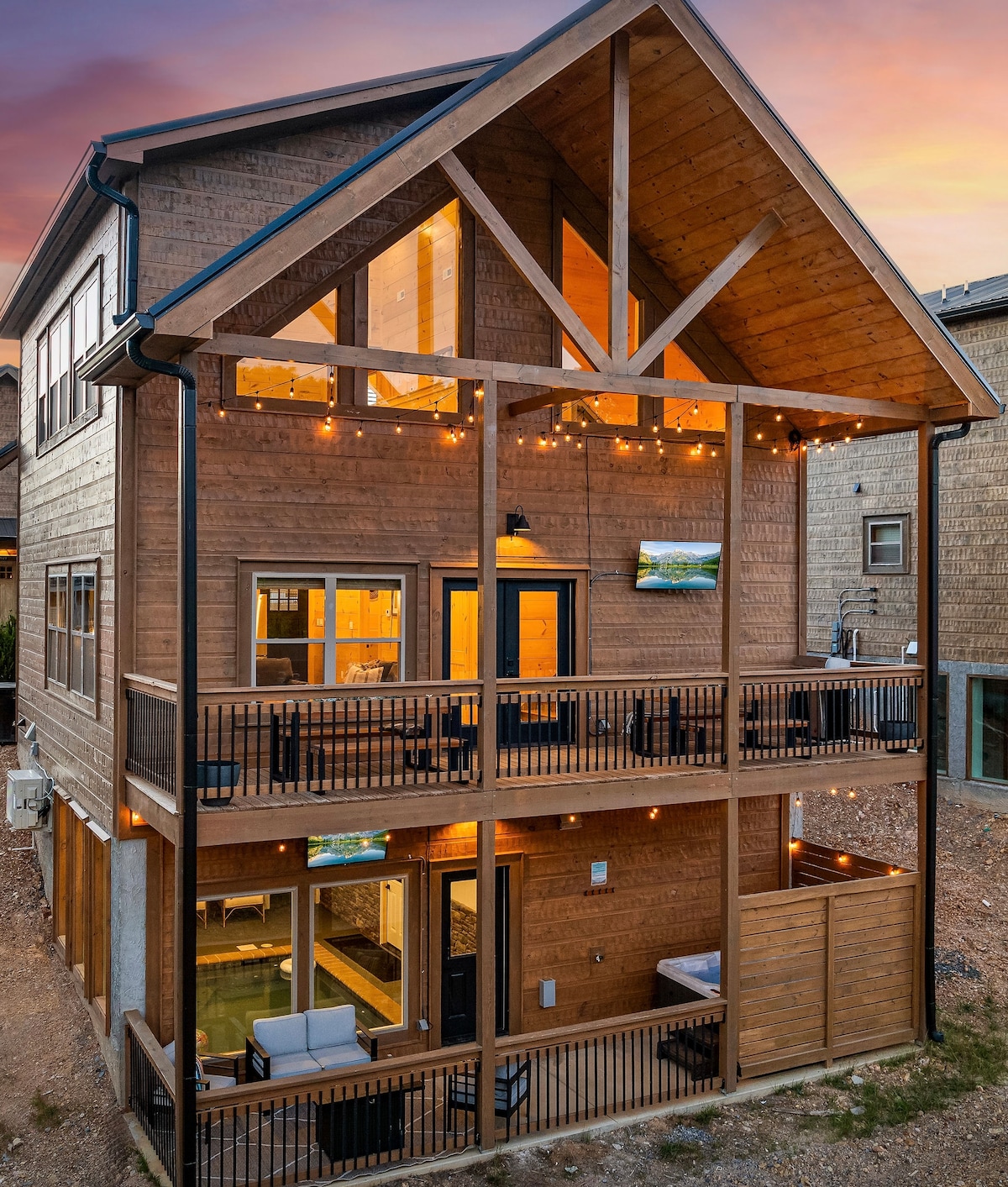 A multi-level cabin is showcased, constructed with a combination of wood and stone. Two outdoor decks are visible, illuminated by string lights. Each level features large windows that allow for ample light. A fire pit and hot tub are situated at the lower deck.