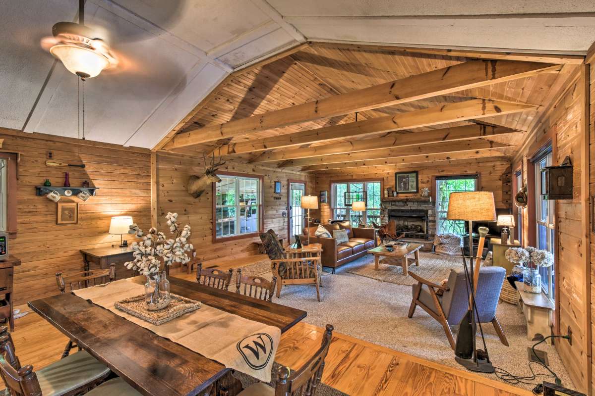 A spacious living area features a wood-paneled ceiling supported by exposed beams. Natural light fills the room through large windows, highlighting comfortable seating arrangements. A dining table is positioned nearby, alongside decorative elements such as a fireplace and stylish lighting fixtures.