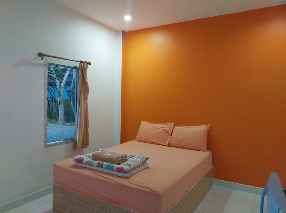 A warm-toned bedroom features an orange accent wall, complemented by a neutral color bed with two pillows. A window offers a glimpse of the outdoors, while neatly arranged towels lie on the bed, enhancing the simple decor.