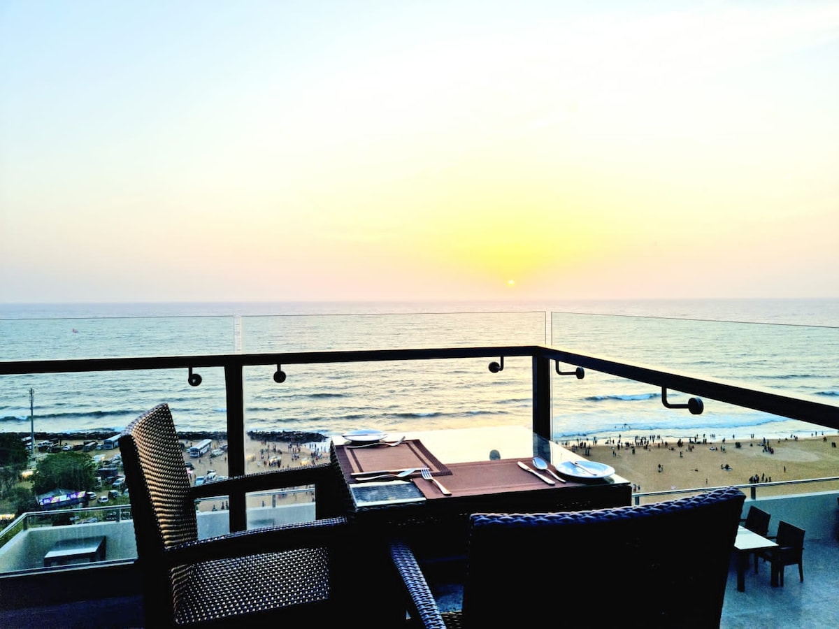 A serene dining area is positioned on a balcony overlooking the ocean. Two woven chairs are arranged around a table set for a meal, while a vibrant sunset casts warm hues on the water. The beach is visible in the distance, with people enjoying the shoreline.