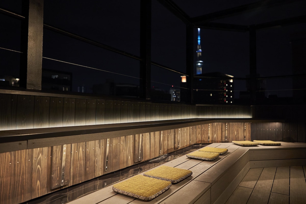 An outdoor private spa space is highlighted by wooden seating and subtle lighting. In the distance, Tokyo Skytree is visible against the night sky, creating a serene backdrop for relaxation.