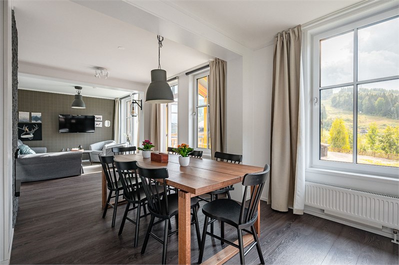 A spacious dining area features a wooden table surrounded by black chairs, with natural light streaming in through large windows. In the background, a living area is visible with a television mounted on the wall and soft furnishings, creating an inviting setting.