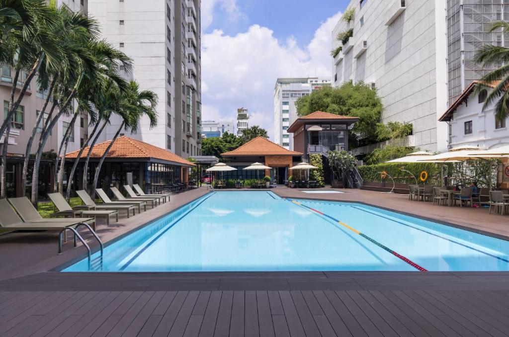 An outdoor swimming pool is surrounded by lounge chairs and palm trees, contributing to a resort-like atmosphere. A shaded cabana with seating is placed nearby, and several umbrellas offer additional comfort. The clear blue water reflects the surrounding buildings and sky.