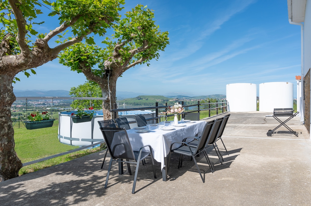 An outdoor dining area is presented, featuring a white table set for six against a backdrop of lush green fields and distant hills. Two decorative trees provide shade, while a swimming pool is visible in the background. The sky is clear, enhancing the scenic view.