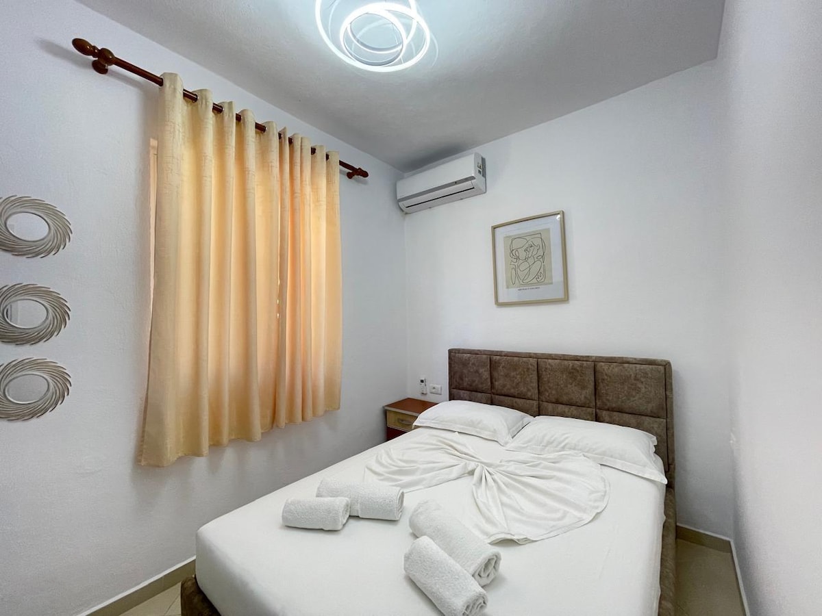 A bright sleeping area features light-colored walls and curtains that softly filter natural light. A comfortable bed is complemented by a decorative towel arrangement. Wall art and decorative mirrors add subtle styling, while an air conditioning unit is positioned above the bed for climate control.