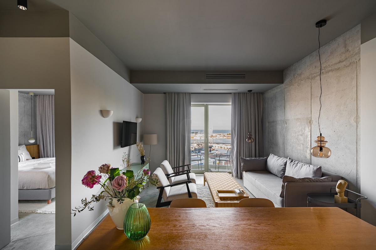 The living area features a light-filled open layout with a comfortable sofa and two chairs positioned around a coffee table. A dining area is visible for four, and large glass doors open to a balcony, offering views of the marina.
