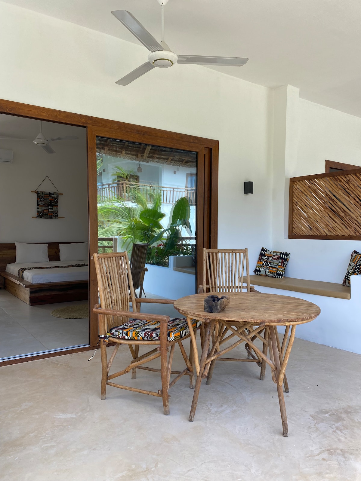 A patio area features a round wooden table with two matching chairs, adorned with colorful fabric. Glass doors lead into the interior space, revealing a sleeping area with decorative artwork and plants visible through the glass.