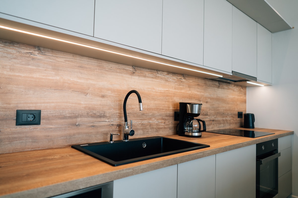 A modern kitchen features sleek cabinetry with a wooden backsplash. A black sink is positioned under angular overhead lighting, accompanied by a coffee maker and kettle. The countertop is made of warm wood, creating a functional and inviting cooking space.