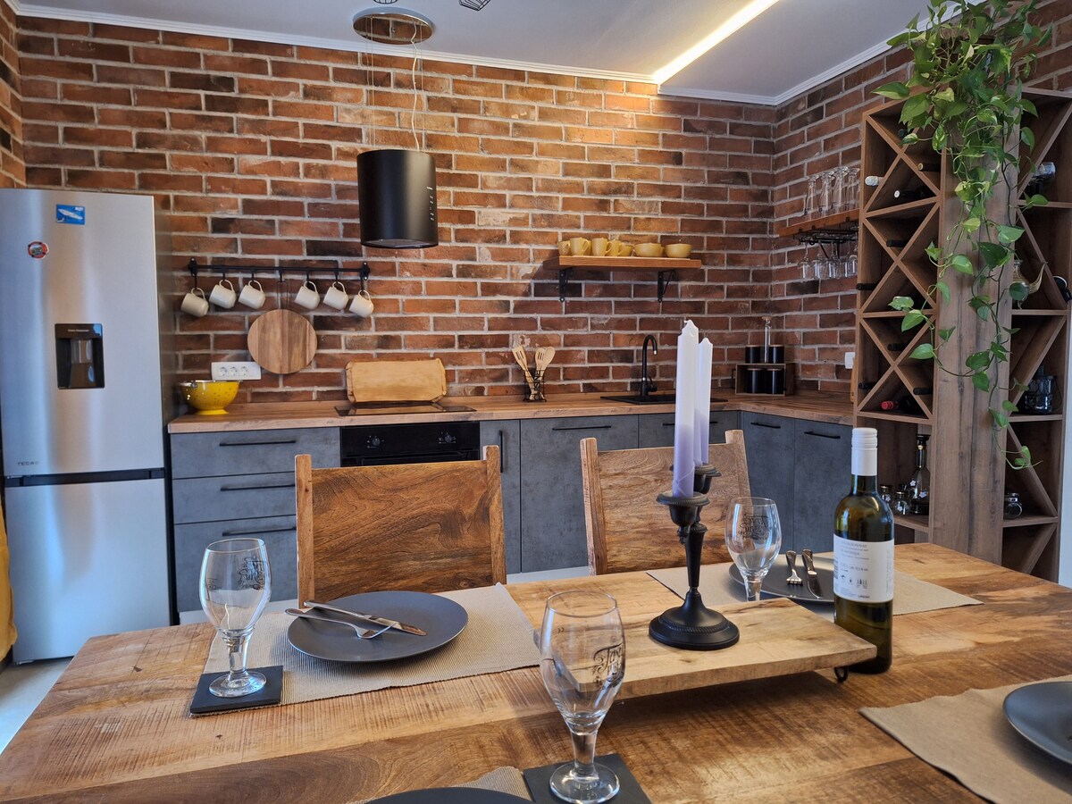 A modern kitchen features exposed brick walls, complemented by a wooden dining table set for a meal. Glassware and plates are arranged neatly, while a lit candle adds a soft ambiance. Functional appliances, including a refrigerator and cabinetry, enhance the space's design.