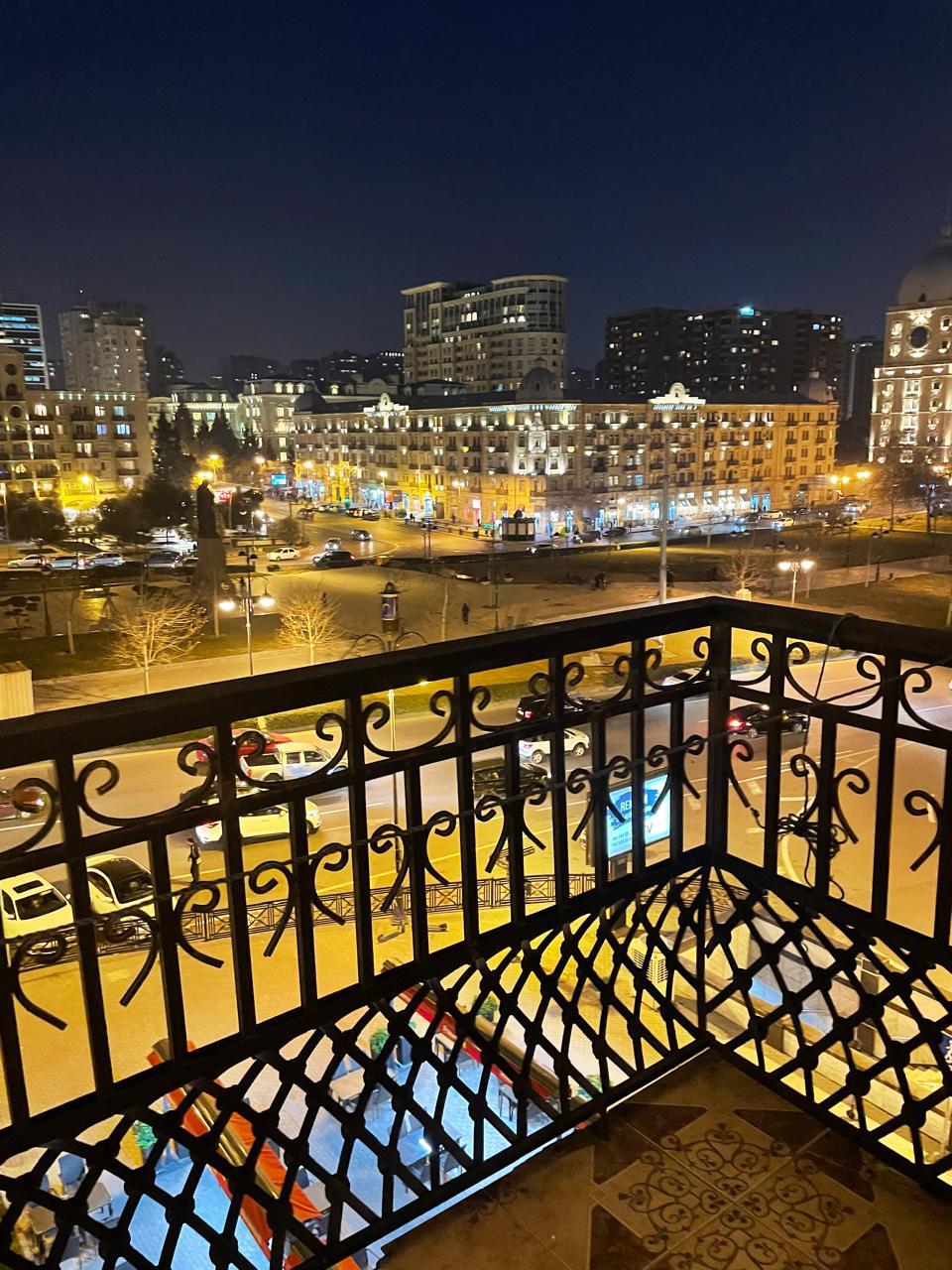 A balcony view captures a vibrant city scene at night, illuminated by soft streetlights. Buildings with architectural details line the street, while vehicles and pedestrians add life to the surroundings. The intricate iron railing frames the scene, creating an inviting outdoor space.