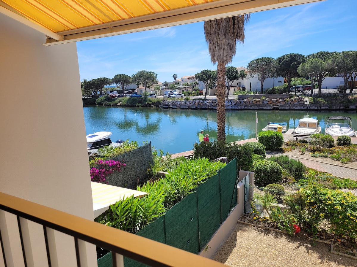 A view from the balcony overlooks a calm marina with boats moored along the waterway. Lush greenery and well-kept flowerbeds frame the scene, while a path leads towards the waterfront. The sky is clear and bright, enhancing the tranquil ambiance of the surrounding area.