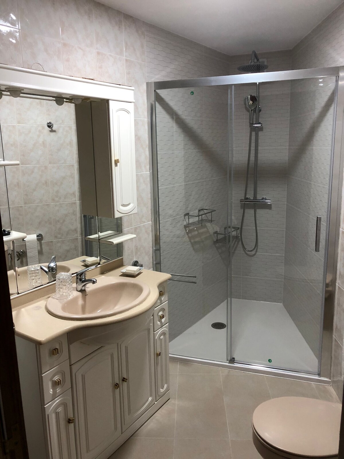A bathroom is shown, featuring a clear glass shower enclosure and a wall-mounted showerhead. A light-colored vanity with a sink is positioned next to a large mirror, reflecting the room's neutral tile design. Soft lighting offers visibility throughout the space.
