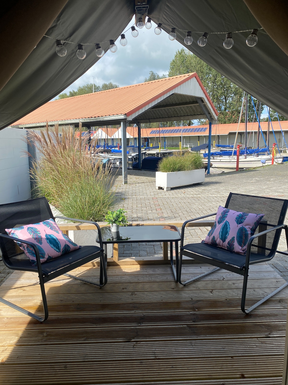 A covered wooden veranda features two black chairs with colorful patterned pillows and a small table. The space offers a view of a marina with docked boats in the background, surrounded by green plants and bright blue skies.