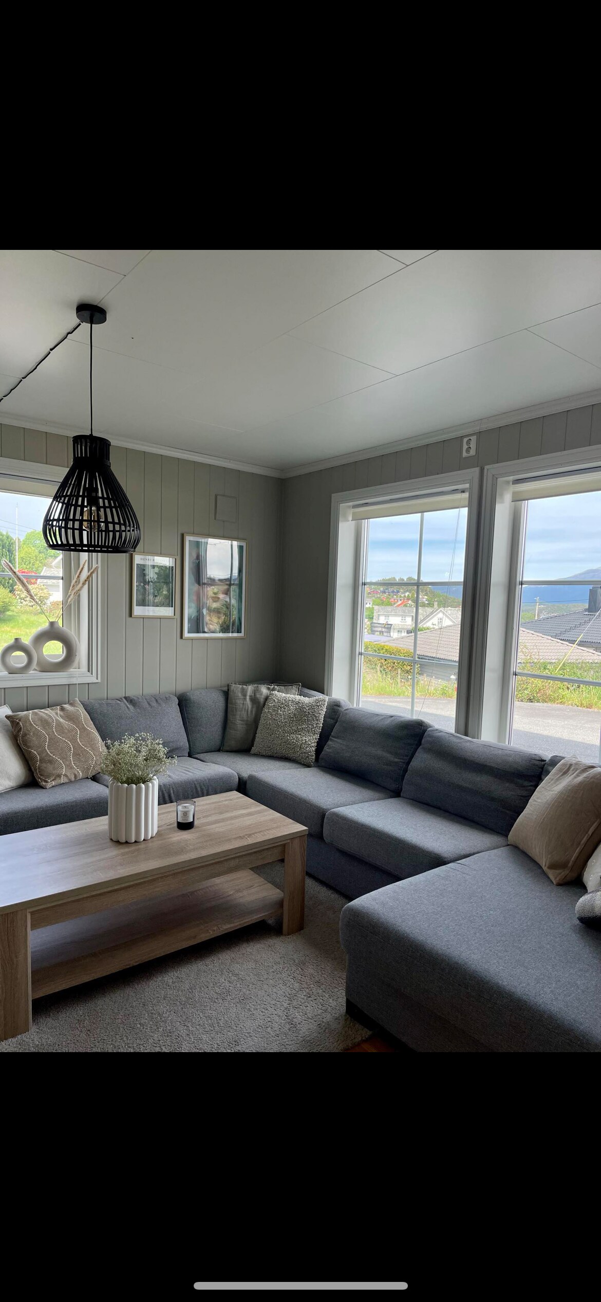 A cozy living area features a large sectional sofa adorned with pillows, positioned near bright windows offering views of the outdoors. A wooden coffee table sits at the center, accompanied by a decorative vase and framed art on the walls, enhancing the inviting ambiance.