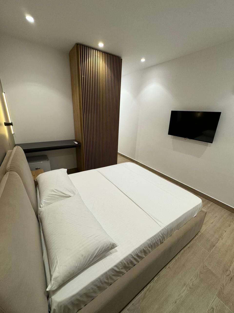 A cozy sleeping area features a spacious bed with crisp white linens, complemented by soft lighting from recessed ceiling fixtures. A flat-screen TV is mounted on the wall, and a wooden accent wall adds a modern touch to the overall decor.