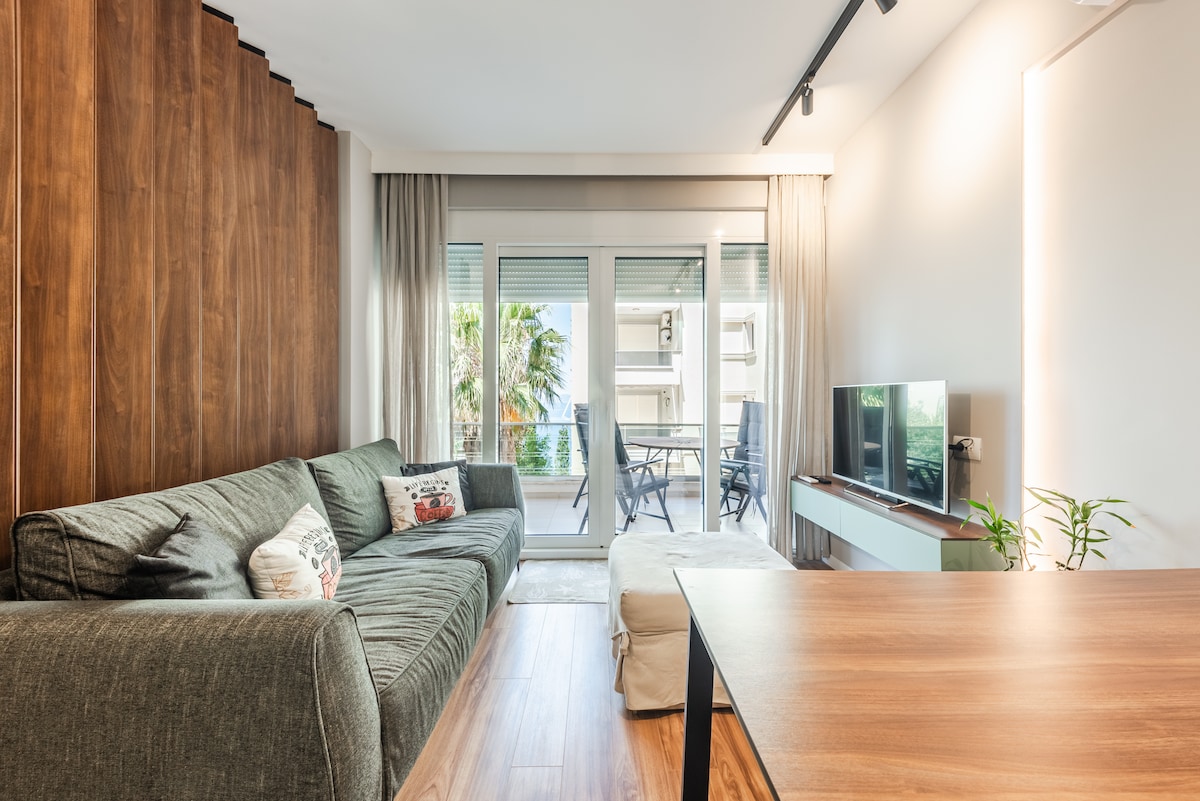 The living room features a comfortable gray couch with decorative pillows, a small ottoman, and a wooden coffee table. Large glass doors lead to a private outdoor area, where a dining table is visible. Natural light fills the space, highlighting the warm wood accents.