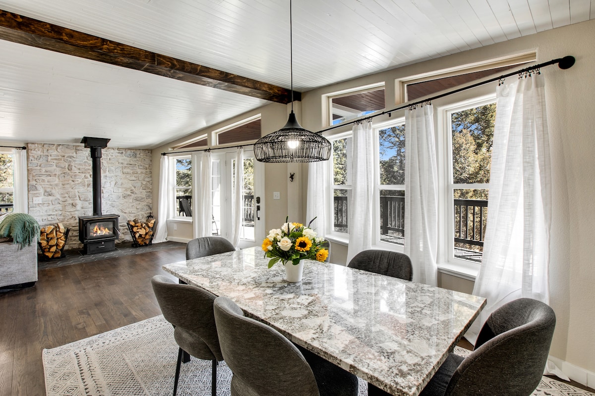 A spacious dining area features a large granite table surrounded by comfortable chairs. Natural light floods the room through large windows, which frame views of the outdoors. A decorative light fixture hangs above the table, and a cozy fireplace adds a touch of warmth to the space.