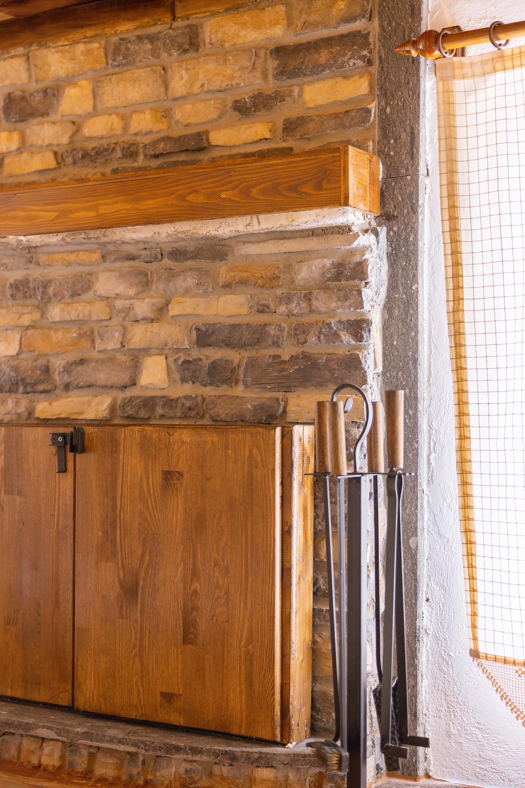 A textured stone wall is complemented by a wooden shelf above and a wooden cabinet beneath. Metal fireplace tools are positioned next to the cabinet, with natural light filtering through a window adorned with a grid-patterned curtain.