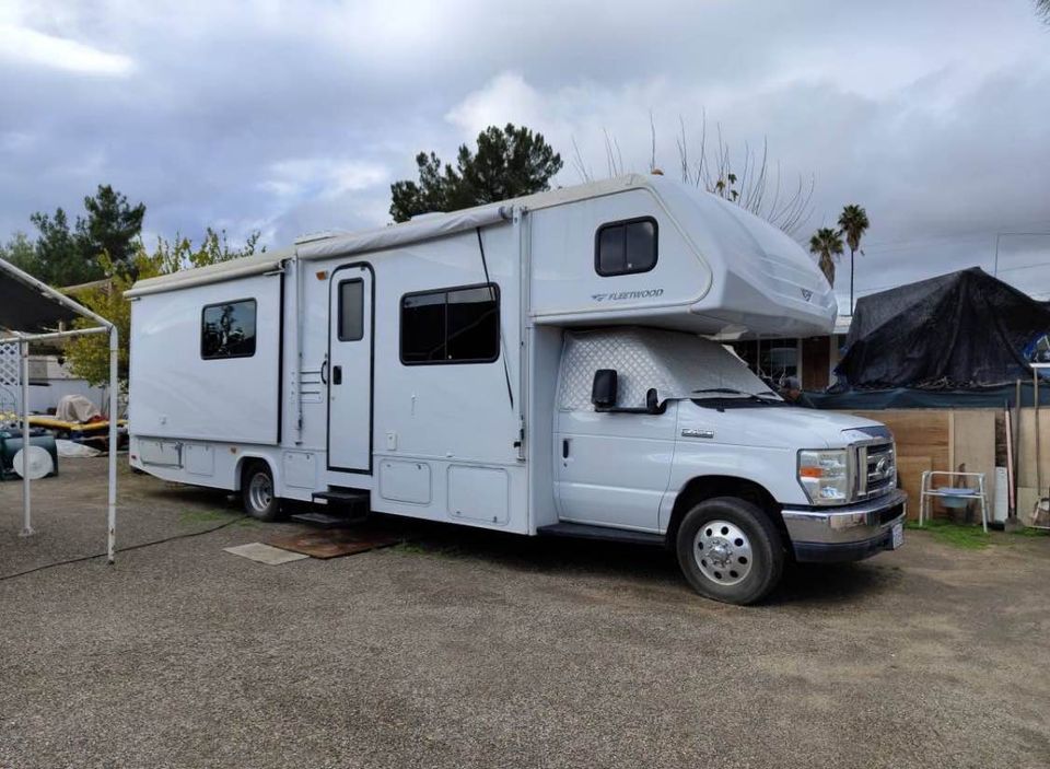 A white recreational vehicle is parked in a gravel area, featuring a large slide-out section on one side. The vehicle displays multiple windows, and the overhead part has an aerodynamic design. Surrounding greenery and distant residential structures are visible in the background.
