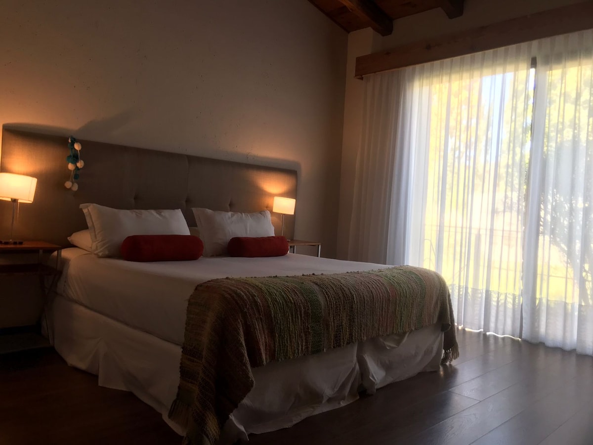 A spacious bed is adorned with soft linens and two decorative pillows. Natural light pours in through a large window covered by sheer curtains, illuminating the warm tones of the room. A bedside table with a lamp provides additional lighting.