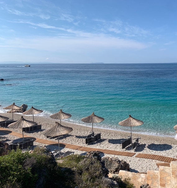 The beach area features several sunbeds shaded by thatched umbrellas, arranged along a sandy shoreline. Clear turquoise water gently laps at the beach, with distant views of the horizon under a blue sky. Natural rocks border the beach, enhancing the coastal scenery.